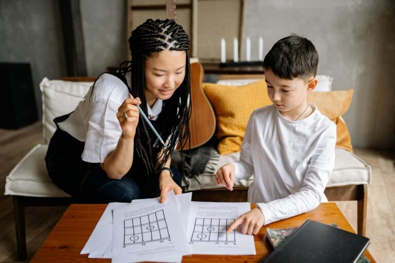 Asian woman and Caucasian boy engaged in a music lesson at home, focusing on paper sheets with a guitar in the background.